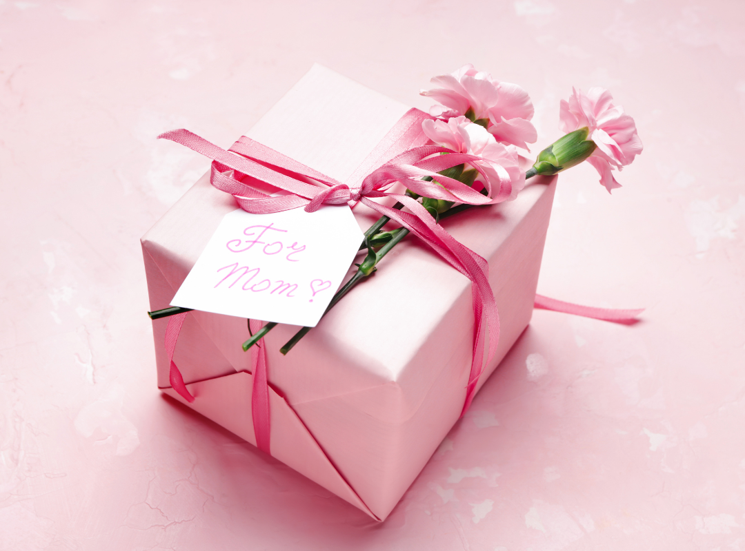 There is a gift wrapped in pink paper and there are pink flowers and a bow on the present. There is a tag that says for Mom with a heart and the table it is on is pink.