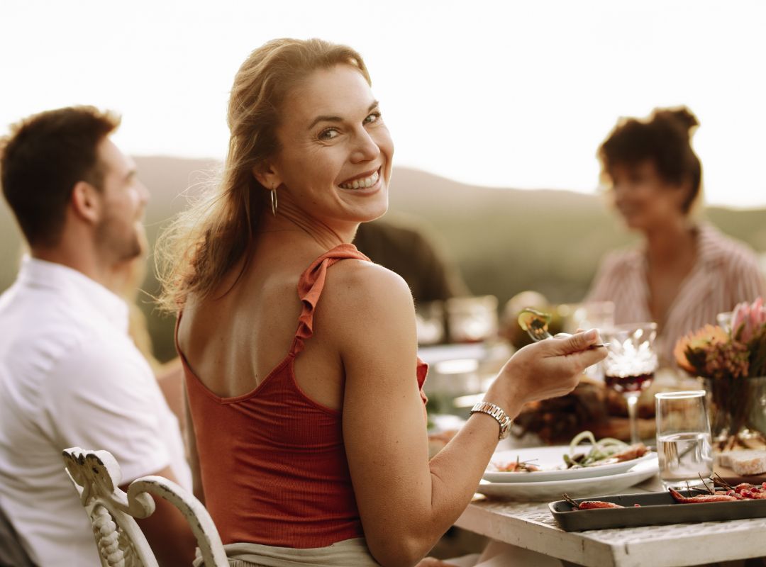 There is a woman in an orange top smiling back at the viewer and she is at a dinner party outside with others.