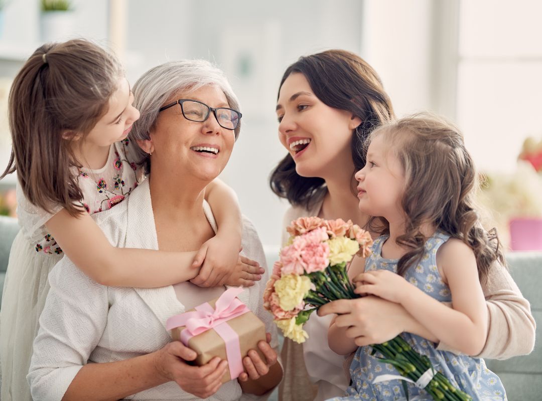 There are two girls in blue and white floral dresses and a grandma with grey hair and a mother with brown hair. They are sitting on a couch and the grandma is holding a present with a pink bow and the Mom is holding pink and white flowers.