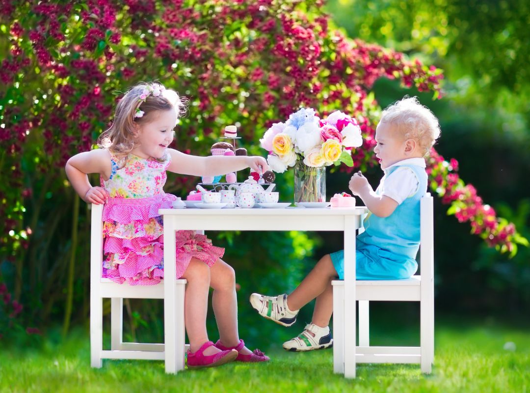 There is a little boy and girl having a tea party outside with red flowers and a white table. They have a tea set and bright clothes on.