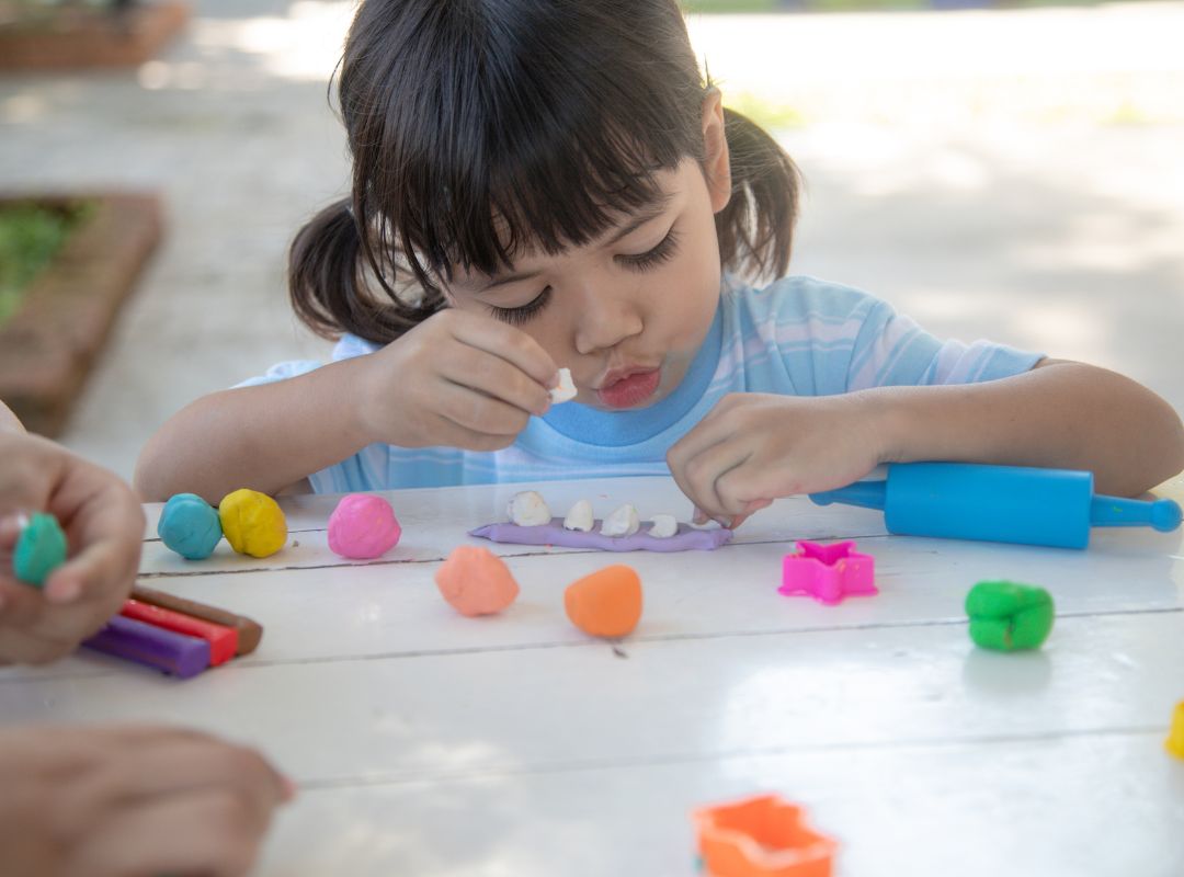 There is a girl playing with playdough outside on a white table.