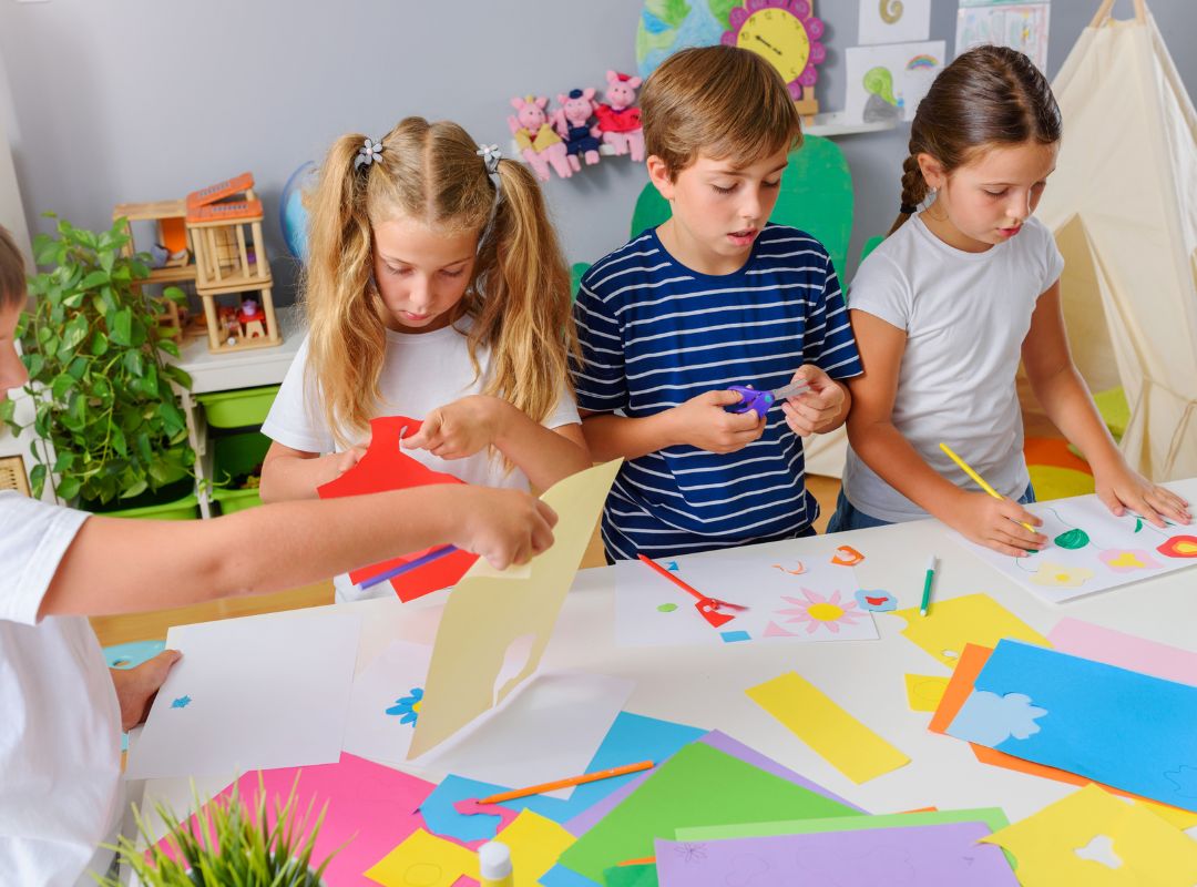 There are kids cutting out paper for crafts on a table.