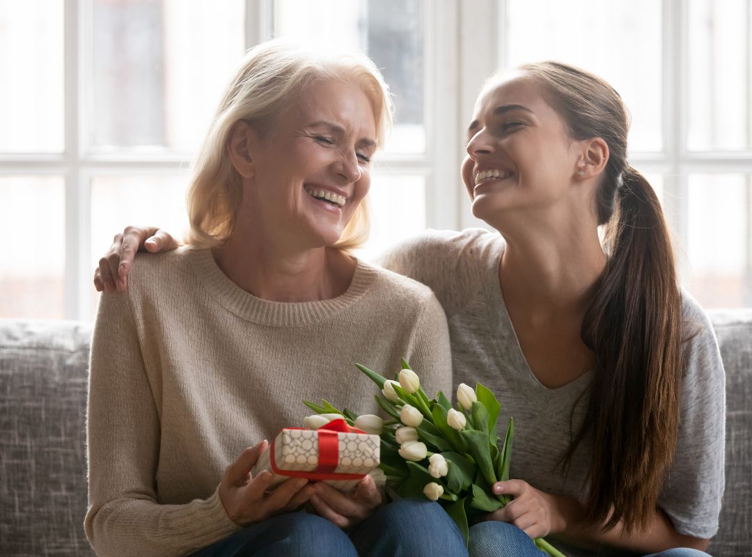 There is a mom and a daughter on a couch and she is receiving flowers and a gift.