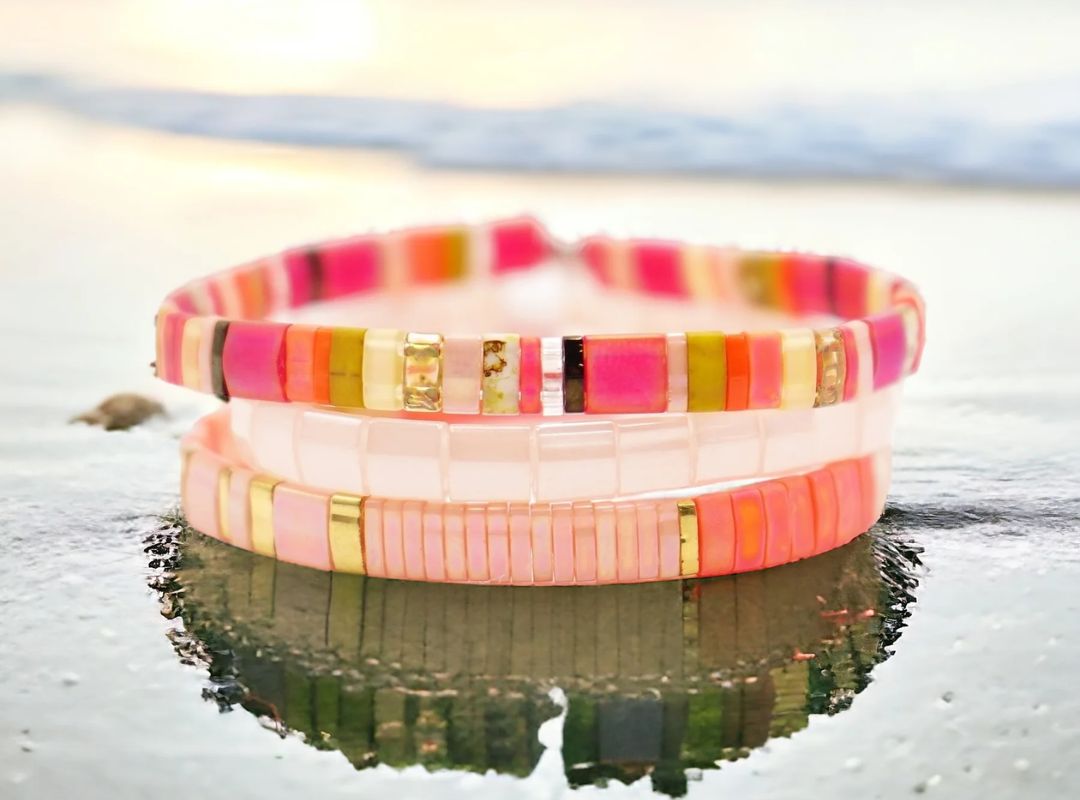 There is a stack of white, gold, and pink beaded bracelets in water on a beach and there is a reflection of the bracelets.