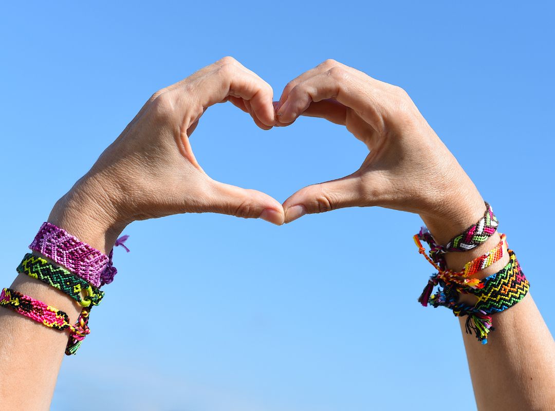 There is a person holding their hands together to make a heart with a blue sky in the background and many bracelets on their wrists.